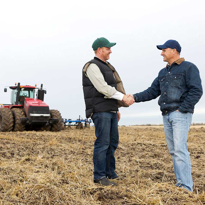 farmer and insurance agent standing in a field shaking hands with a tractor in the background